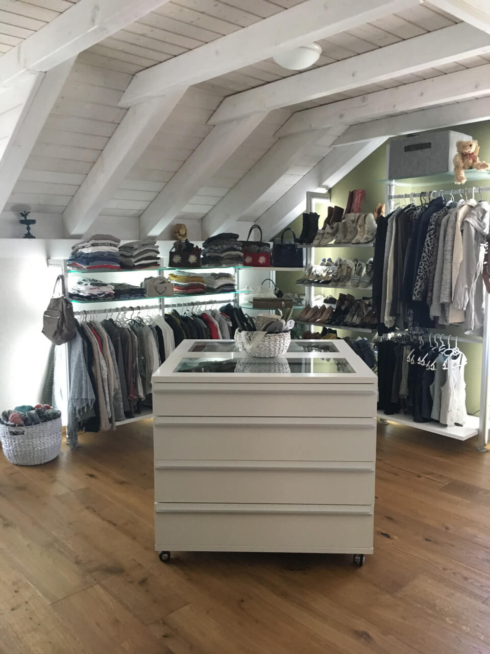 Großzügiges Schranksystem für Dachschrägen mit Glasböden, Kleiderstangen und zentraler Kommode auf Rollen im offenen Ankleideraum